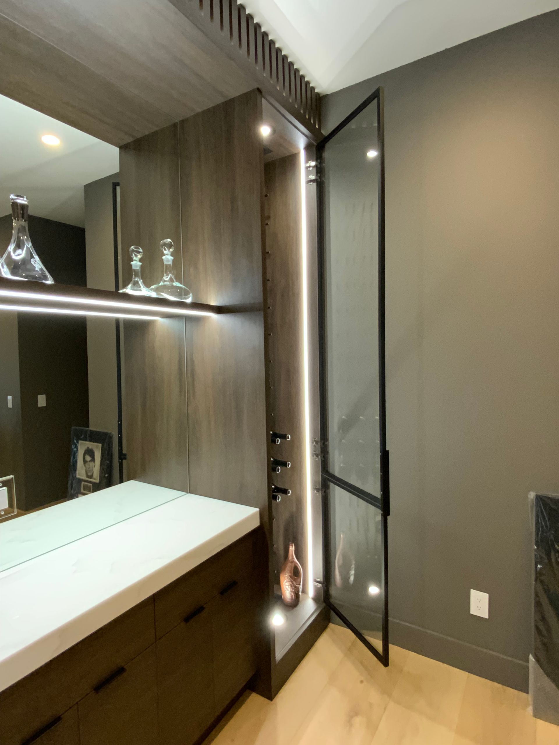 Dark wood built-in bar with a glass door. Features shelves with liquor bottles, led lighting, and a white countertop.