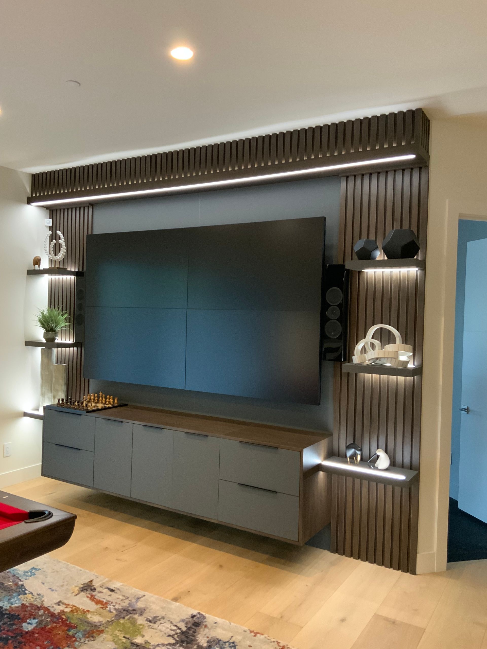Modern media wall with gray TV and floating cabinets, wood paneling, shelves, and LED lighting.