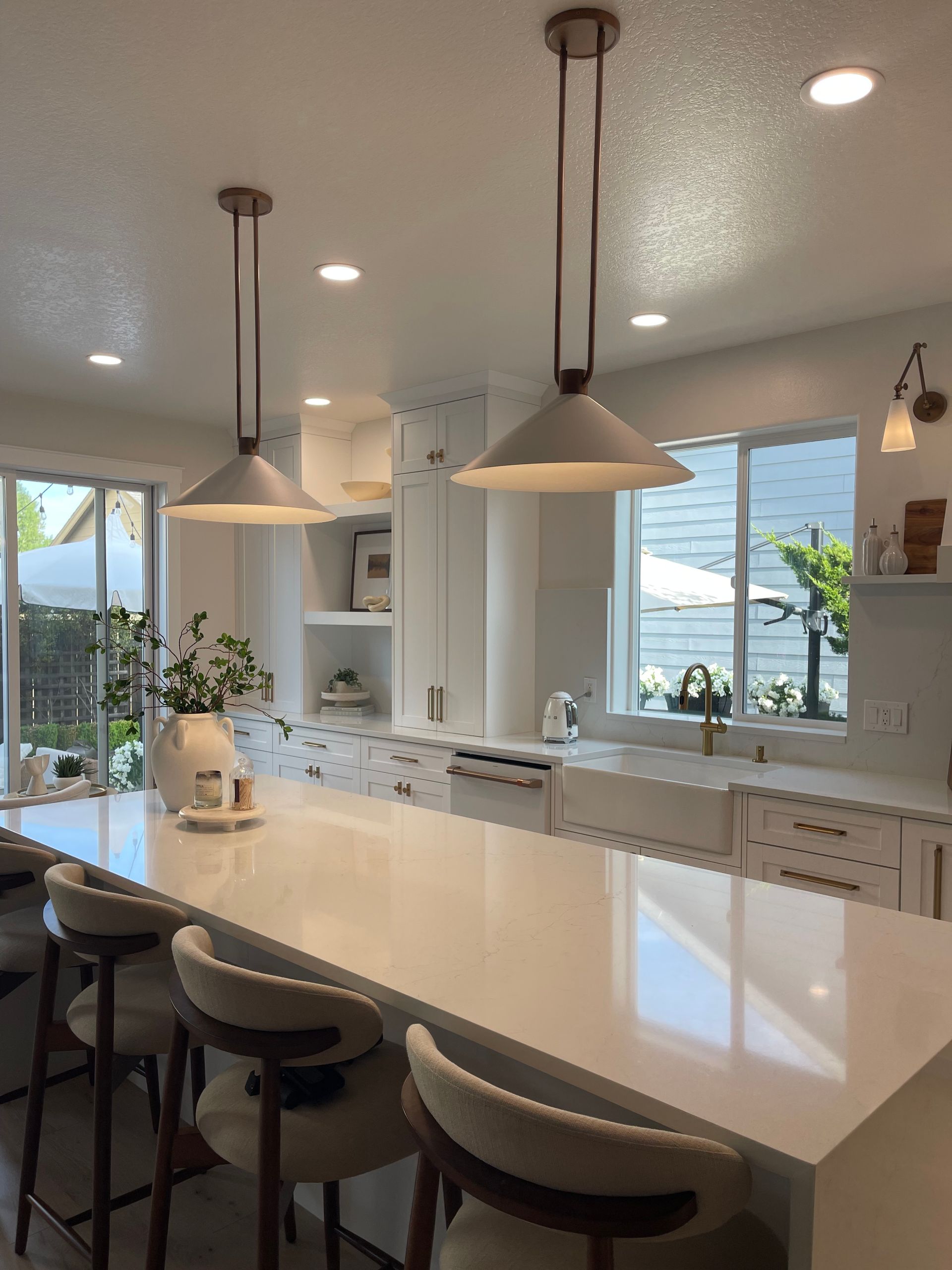 Bright kitchen with a large white island, pendant lights, stools, and windows overlooking greenery.