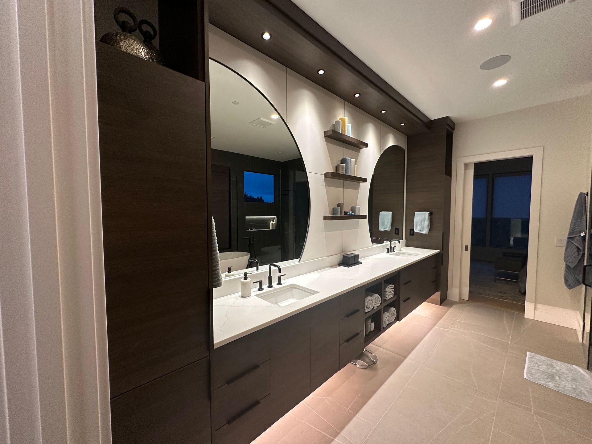 Dark-wood bathroom with double vanity, large round mirrors, and neutral floor tiles. Doorway leads to a view outside.