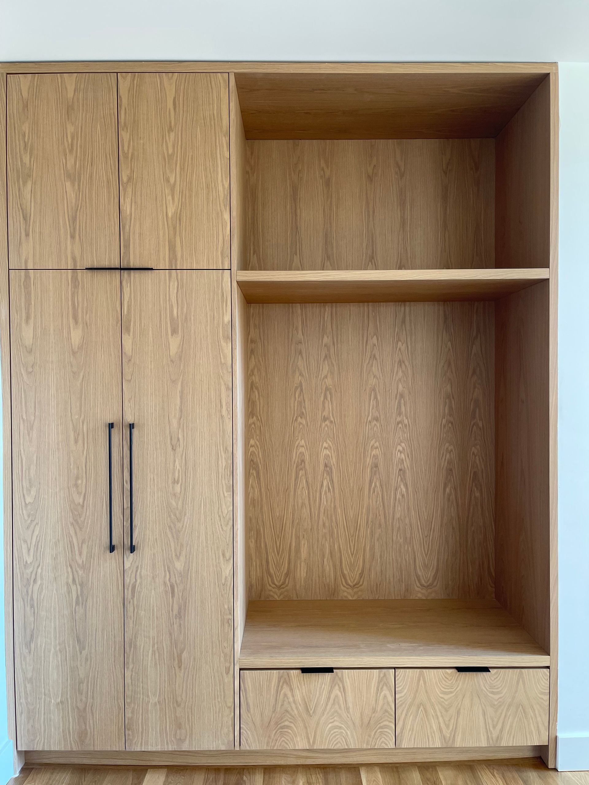 Wooden built-in storage cabinet with doors, shelves, and drawer. Light wood tone.
