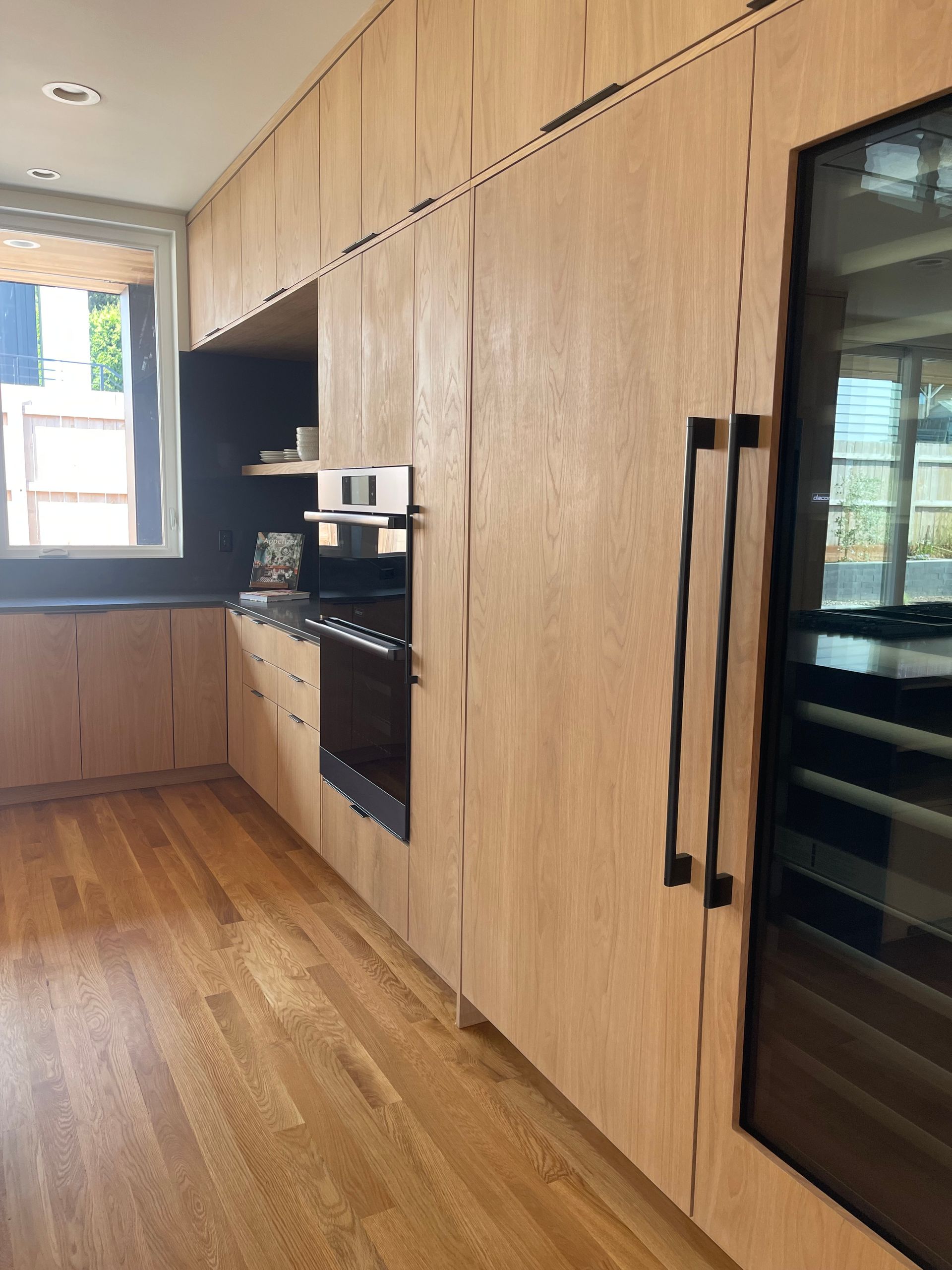 Light wood kitchen with built-in appliances and black handles. Natural light from a window.