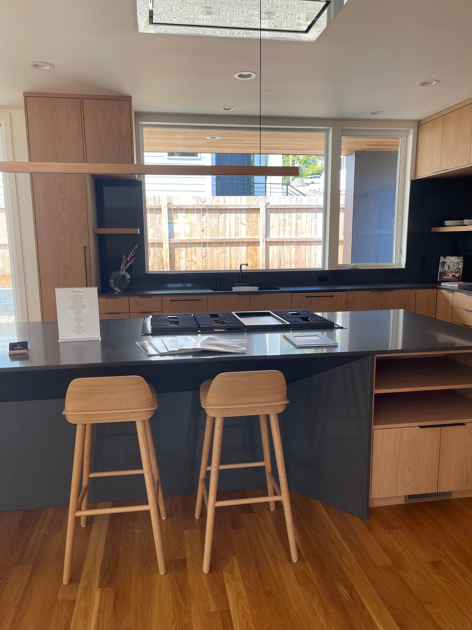Kitchen with island, two wooden stools, and a large window overlooking a fenced backyard.