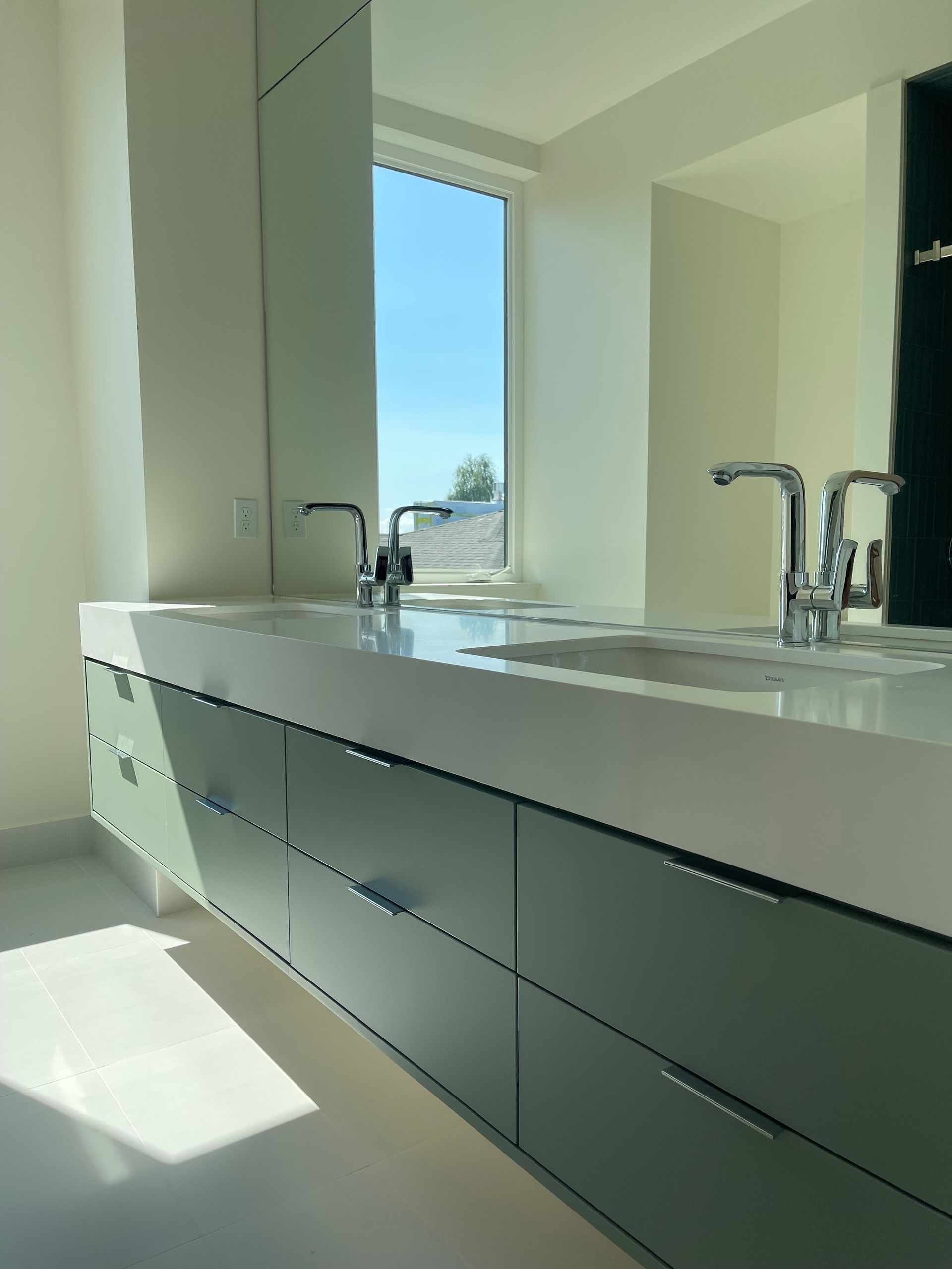 Modern bathroom with light green cabinets, double sinks, and a window with a clear blue sky.