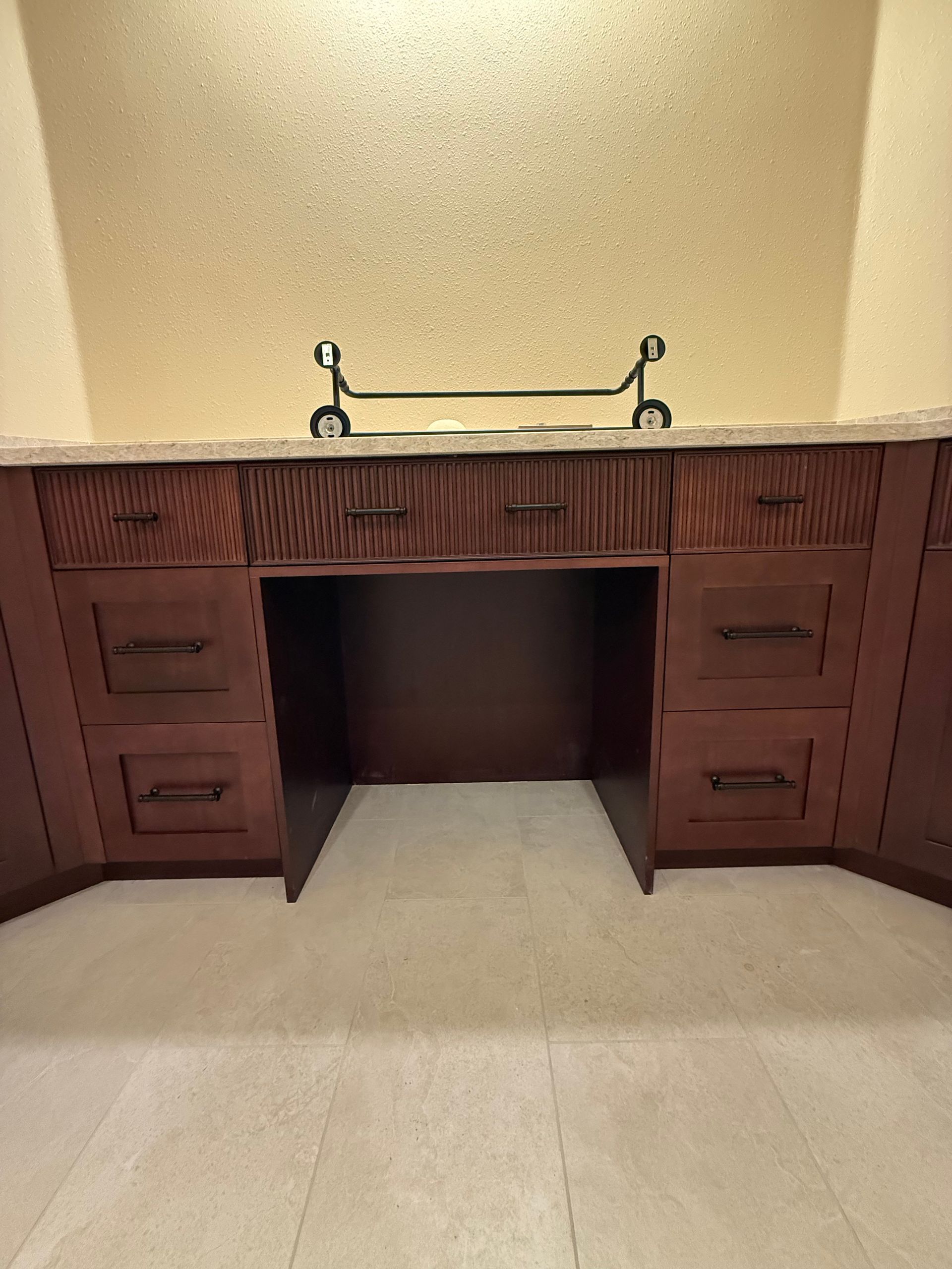 Dark wood desk with drawers, open center for chair, topped with a counter and decorative bar, set against a textured wall.