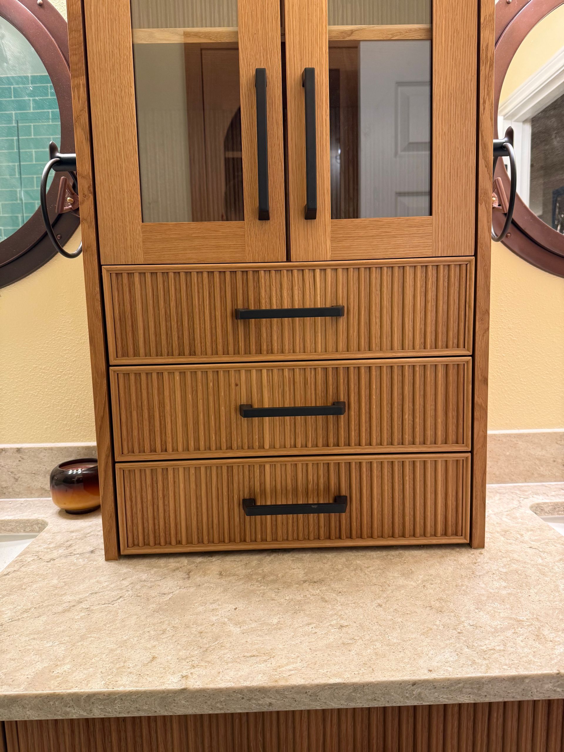 Wooden bathroom cabinet with glass doors and three drawers, set against a bathroom countertop.