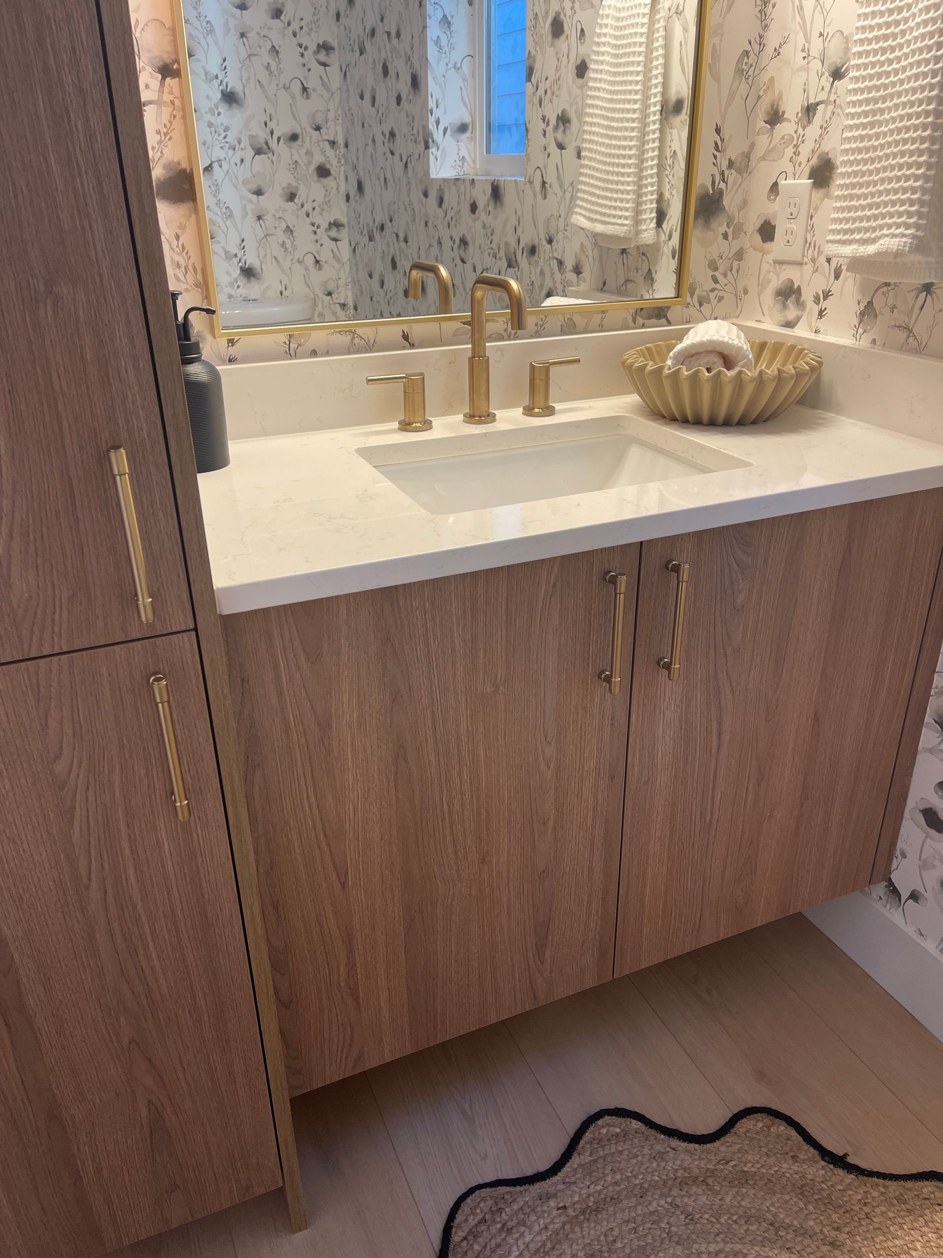 Bathroom with light wood vanity, gold fixtures, white countertop, and patterned wallpaper pro-techwoodwoorking.