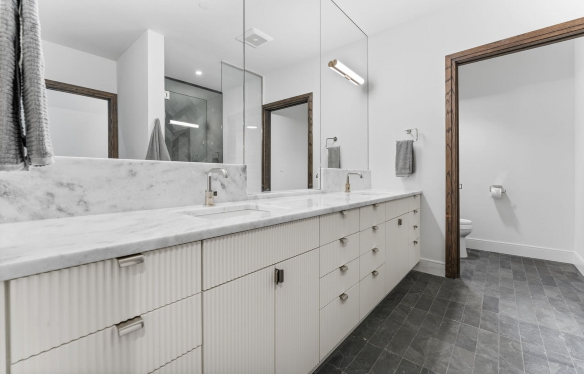 Modern white bathroom with marble countertop and dark gray floor, featuring a double vanity and doorway.