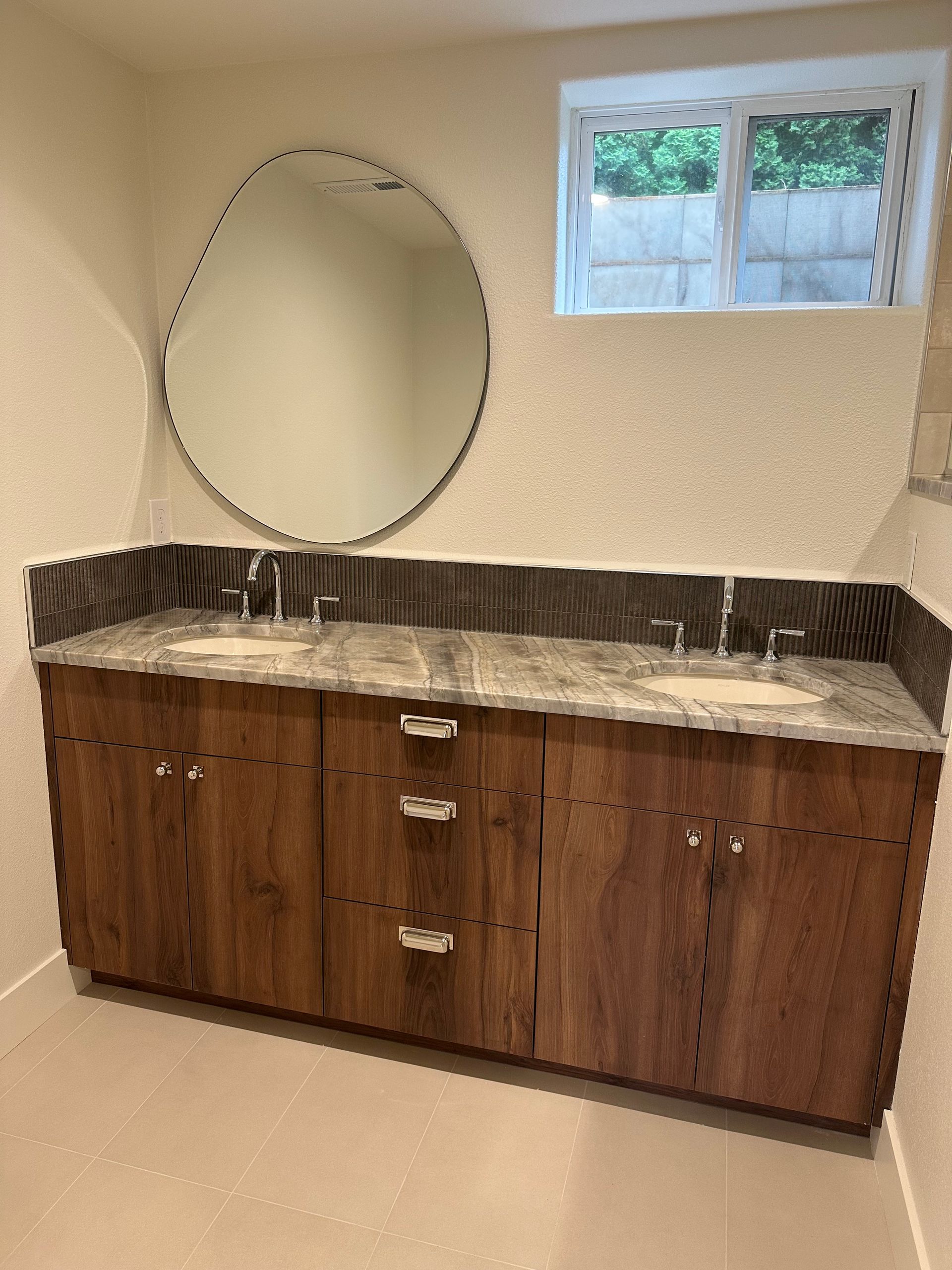 Bathroom with wood double vanity, marble countertop, round mirror, and small window.