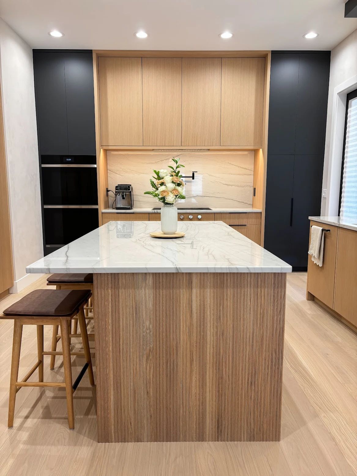 Modern kitchen with a light wood island and cabinets, marble countertop, black accent cabinets, and wood stools.