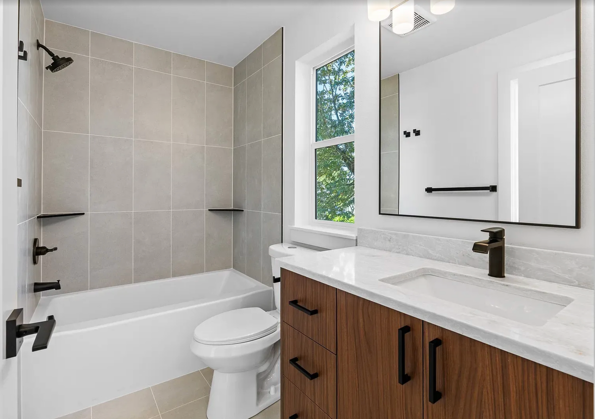 Modern bathroom with a bathtub, toilet, vanity, and window. Light gray tile, wood vanity, black fixtures.