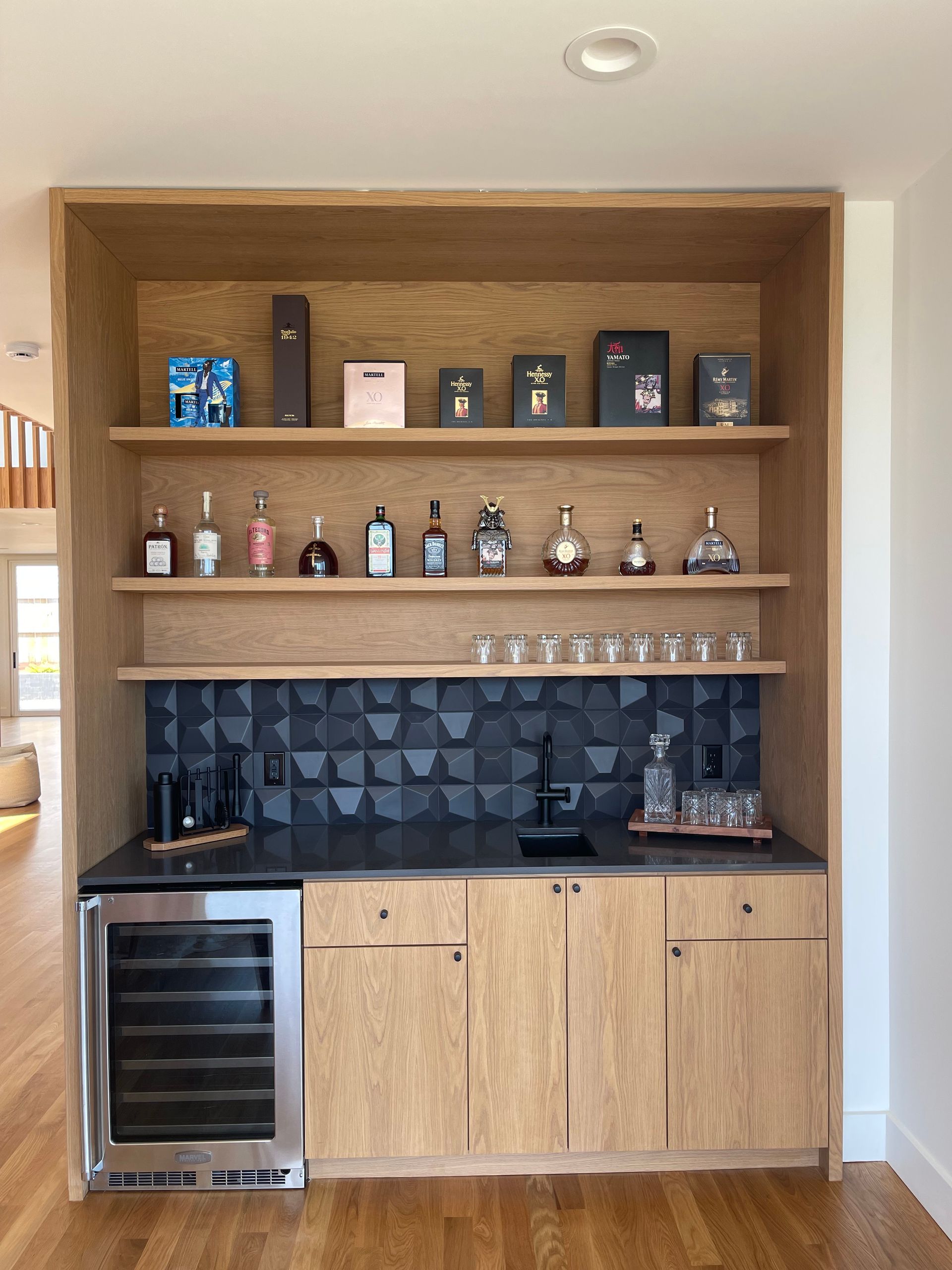Built-in oak home bar with liquor bottles, glassware, a wine fridge, and black backsplash.
