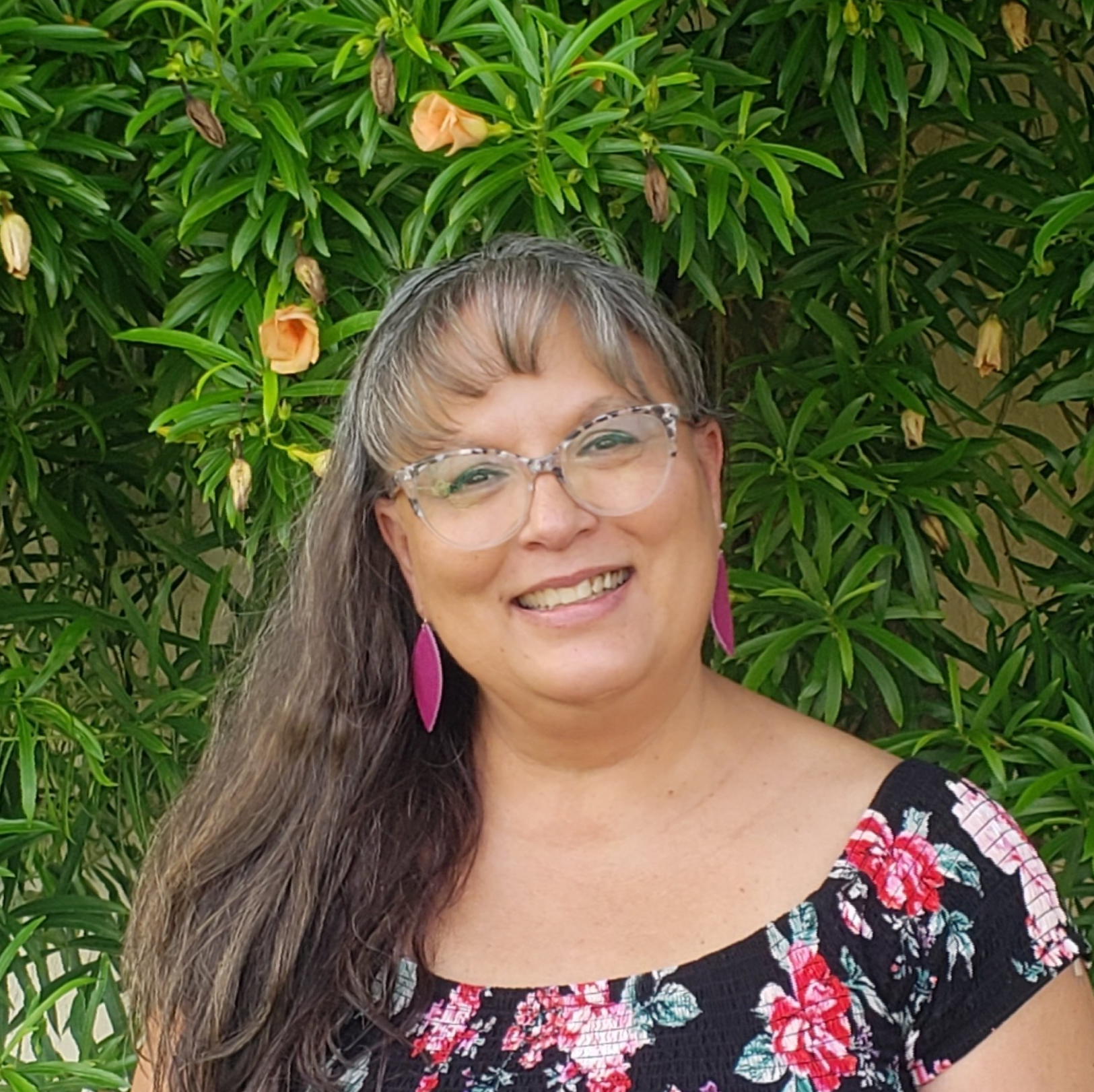 A woman wearing glasses and a floral top is smiling in front of a tree.