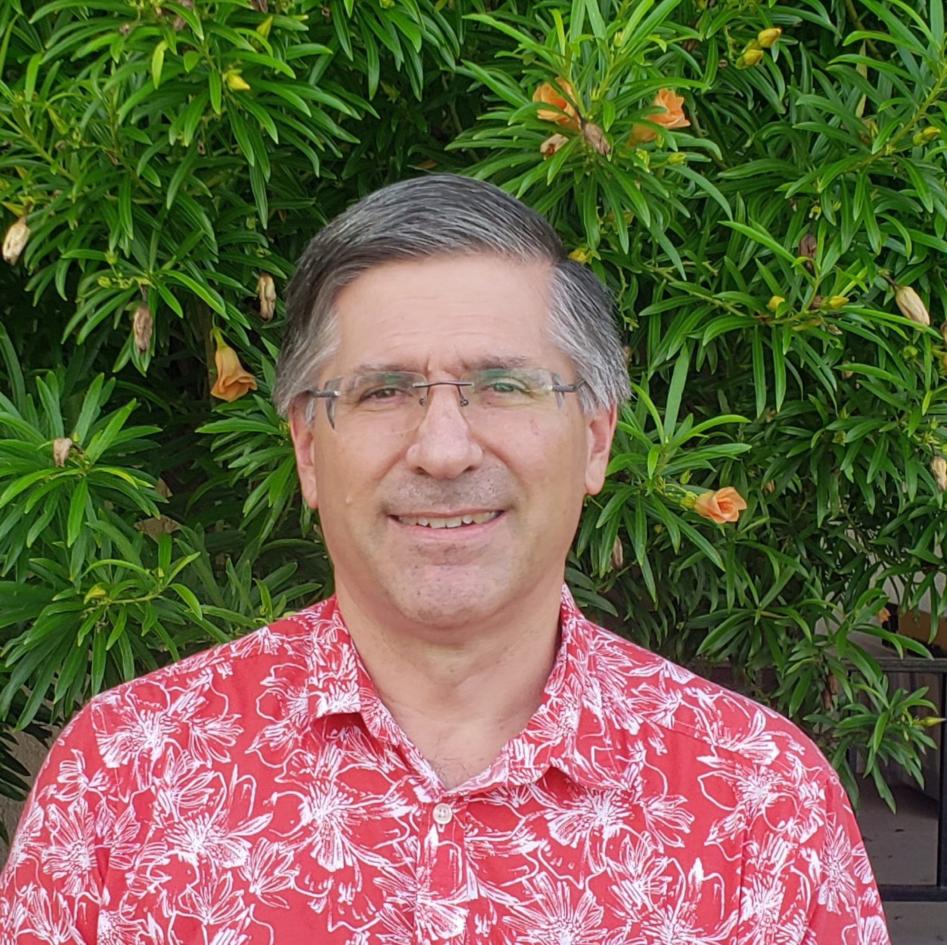 A man wearing glasses and a red shirt is smiling in front of a bush.