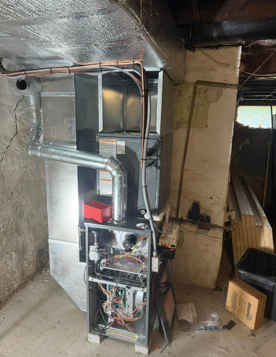 A furnace unit with exposed internal wiring, metal ductwork, and copper pipes in an unfinished basement.