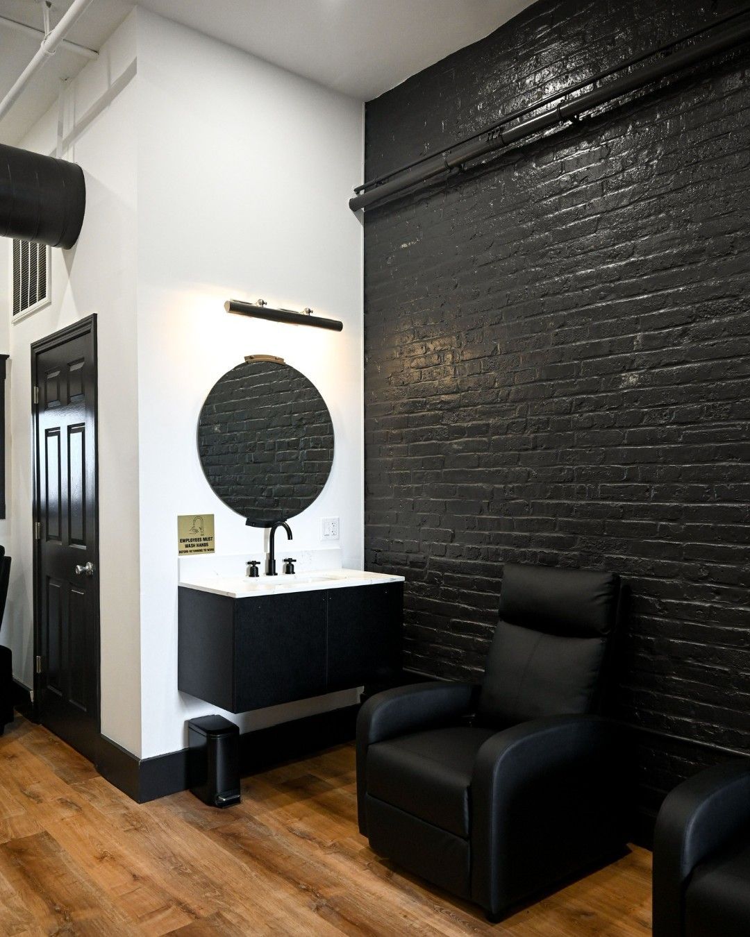 Boston Beautox med spa treatment room- Black and white bathroom with black brick wall, vanity, and recliner chair on wood floor.