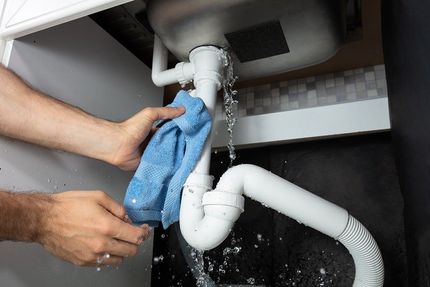 Person holding a blue cloth under a leaking sink pipe in a white cabinet. Water is spraying.