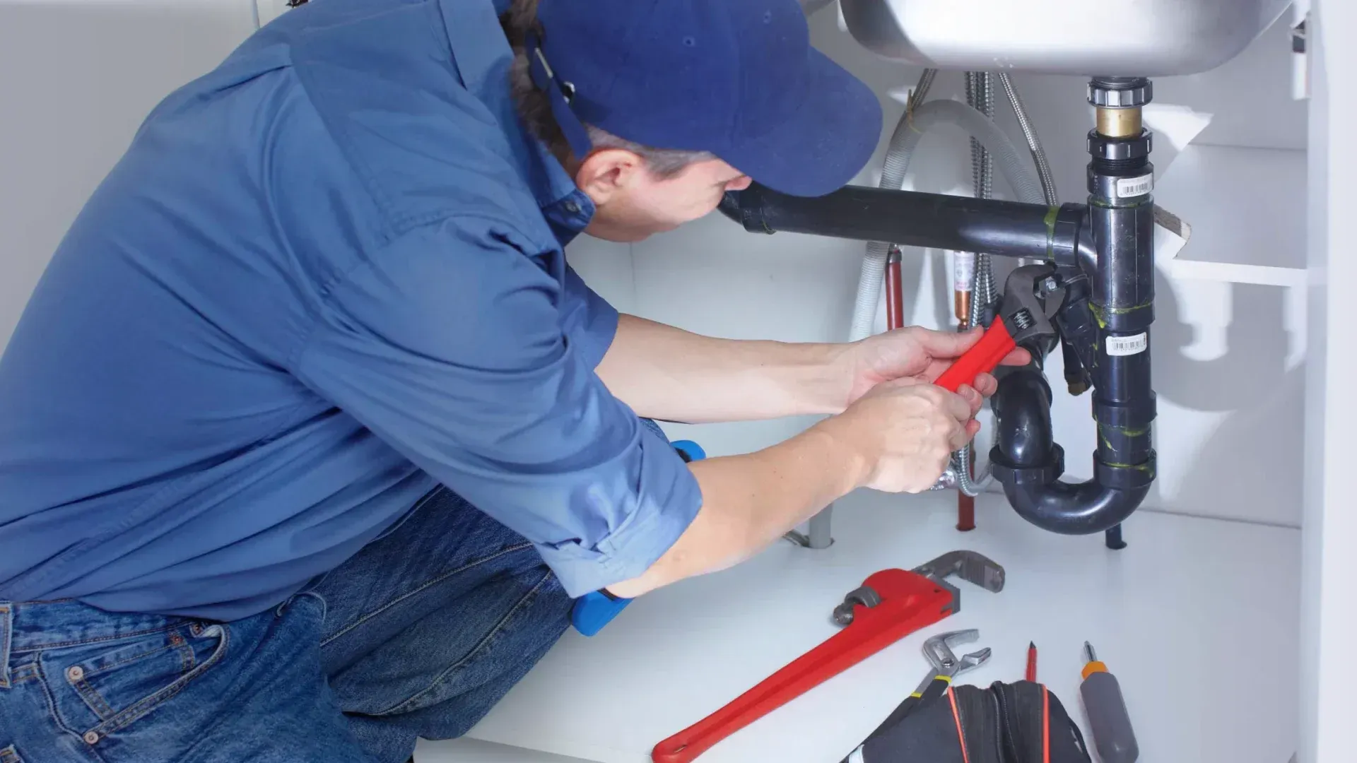 Plumber working under a sink, using a wrench. Blue shirt, hat, black pipe, white cabinet.