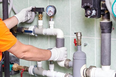 Plumber in orange shirt and gloves, working on a complex system of pipes and valves against a tiled wall.
