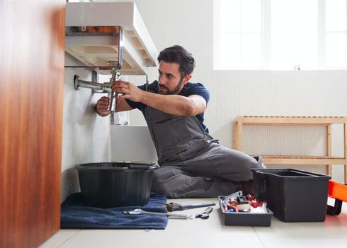 Plumber fixing pipes under a bathroom sink, using tools. Black basin catches drips.