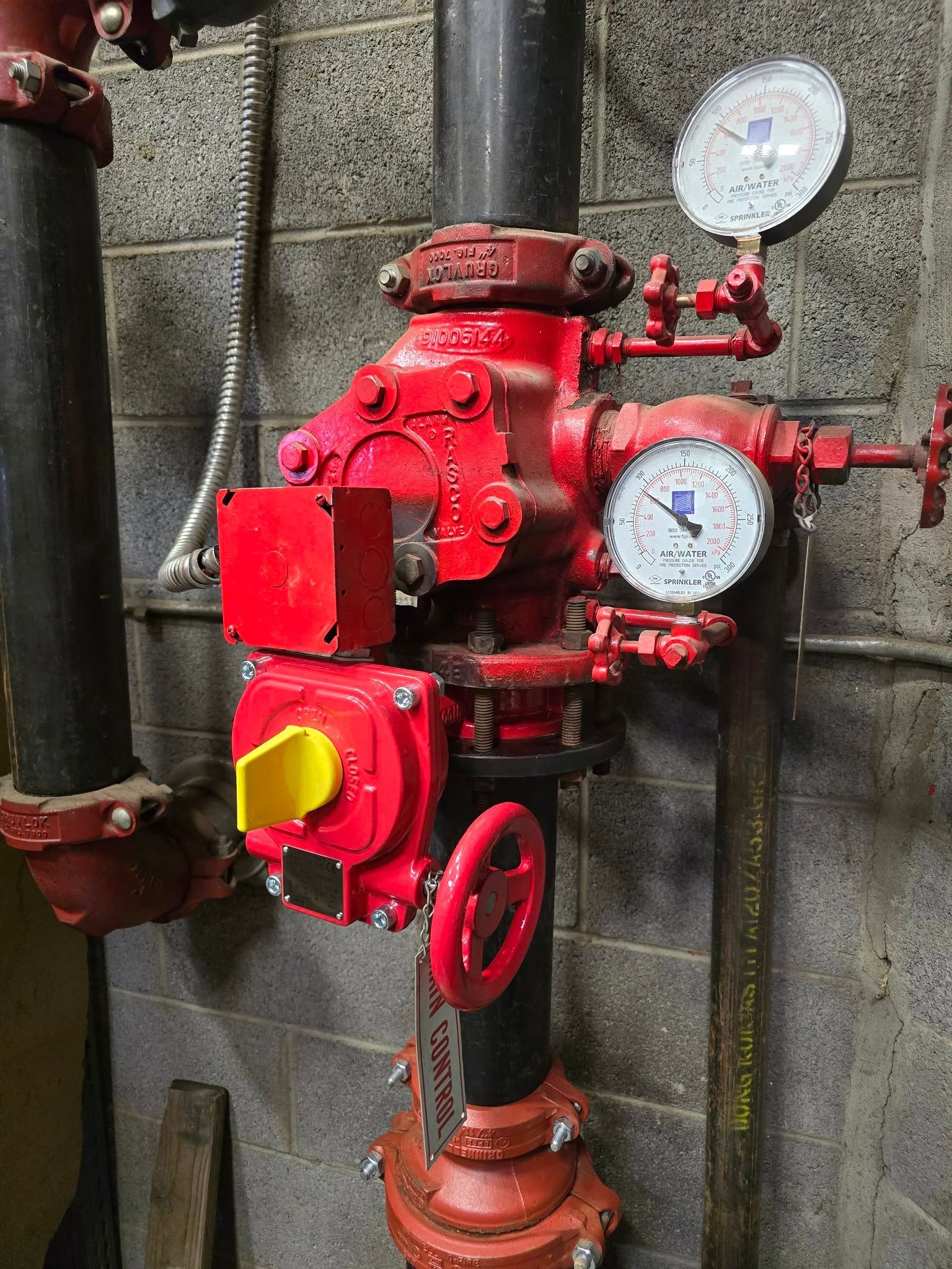 Red fire sprinkler system control valve with gauges, black pipe, and gray block wall.