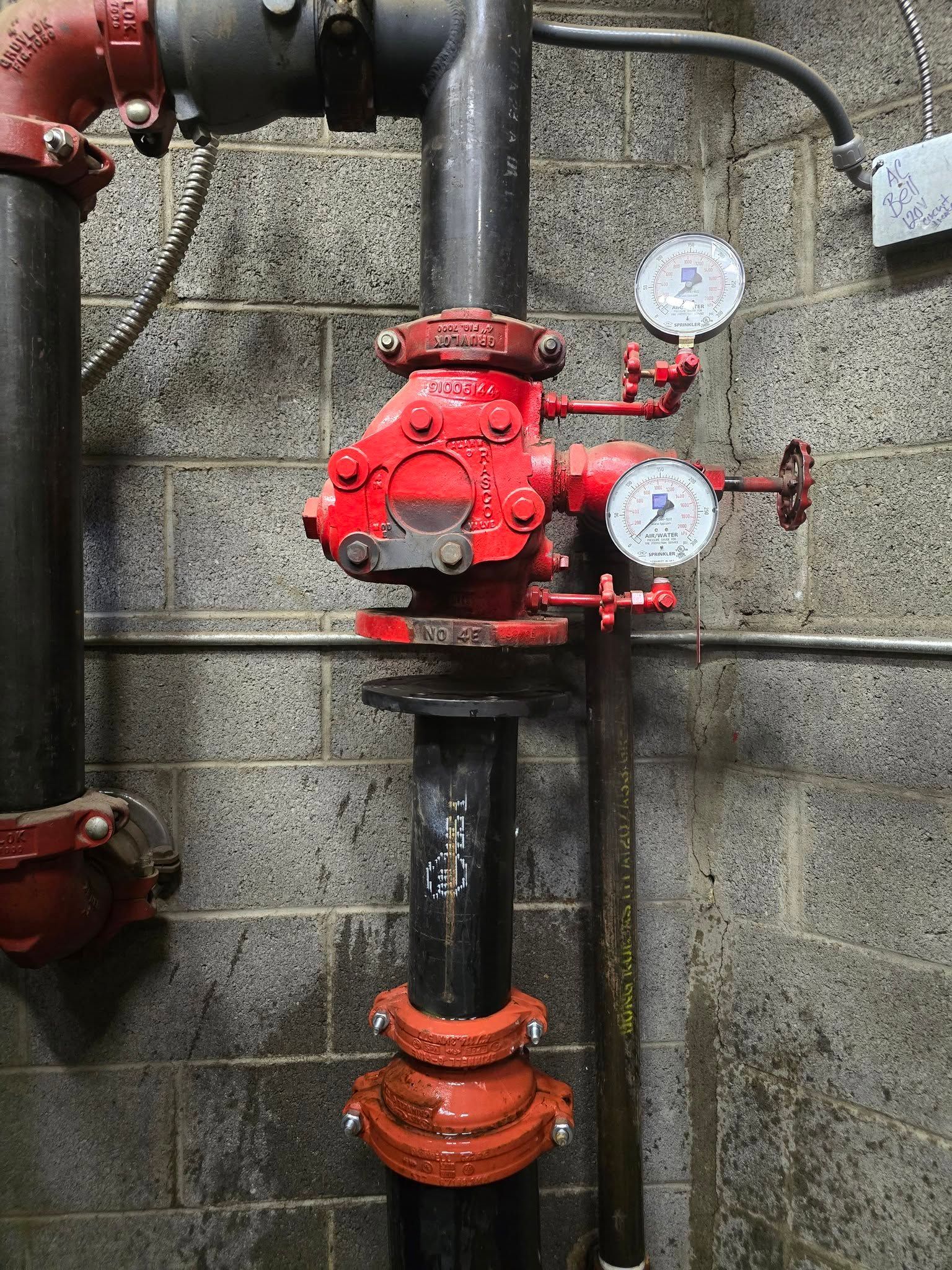 Red fire sprinkler system components on a black pipe against a concrete block wall.