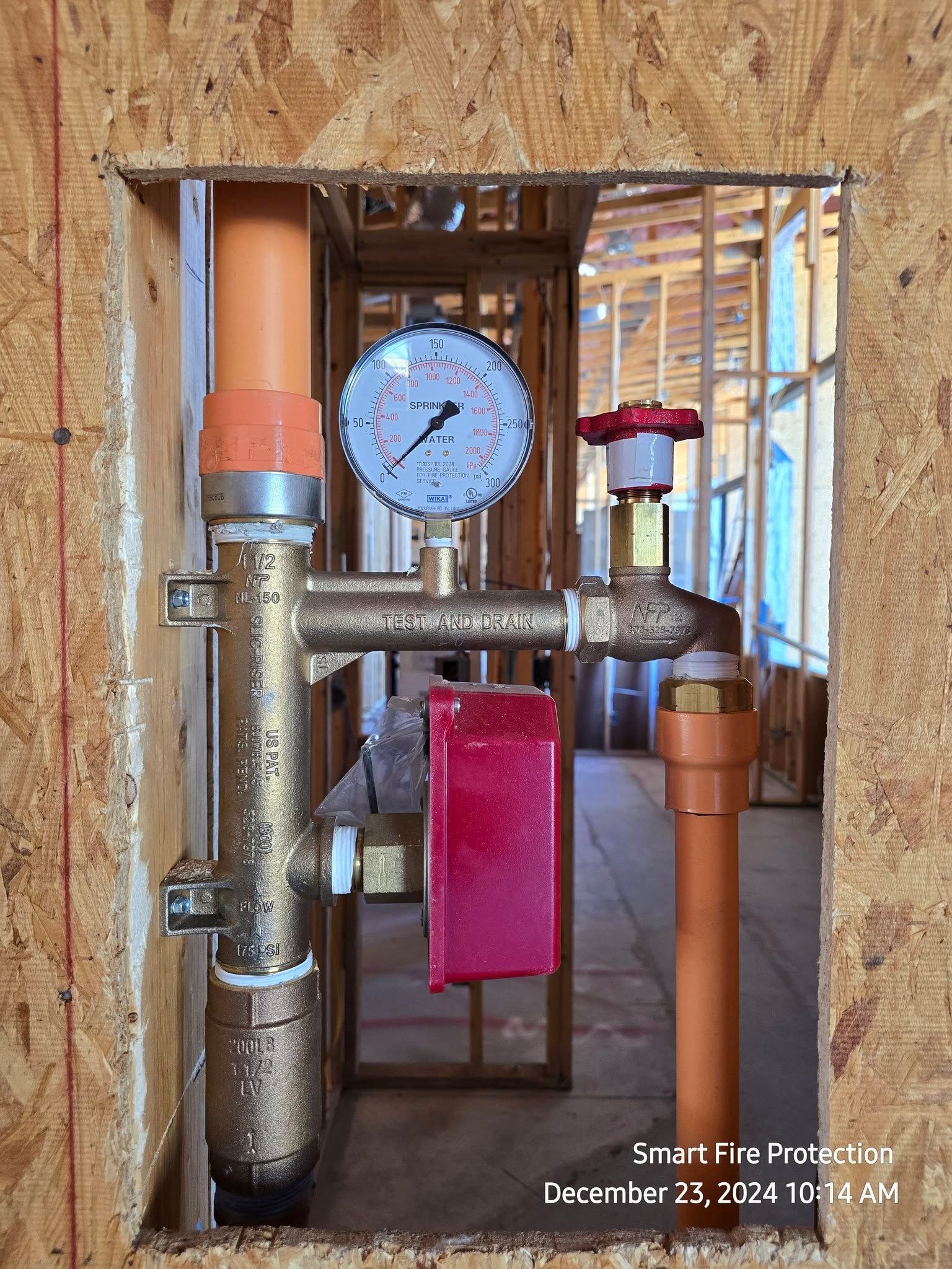 Fire sprinkler system components installed in a wall opening. Brass pipes, pressure gauge, red alarm, orange pipes.