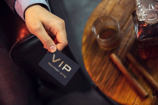 A person is holding a vip card in front of a table with a glass of whiskey and cigars.