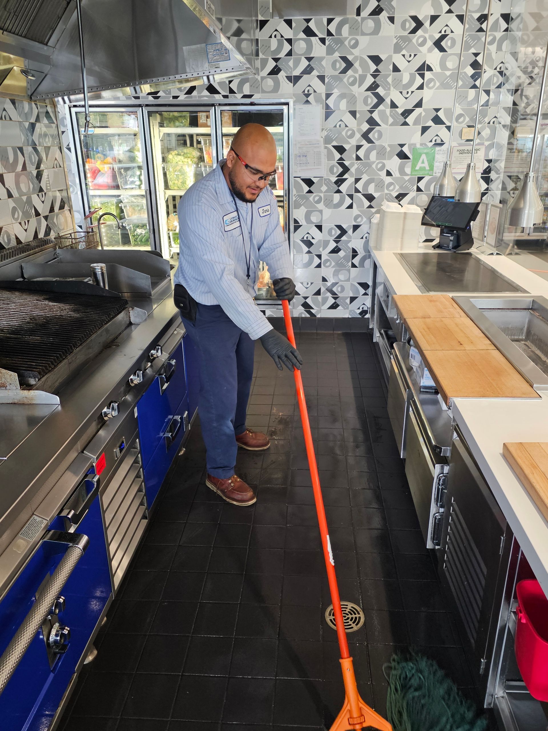 Man mopping a restaurant kitchen floor. He is wearing gloves and a light blue shirt.