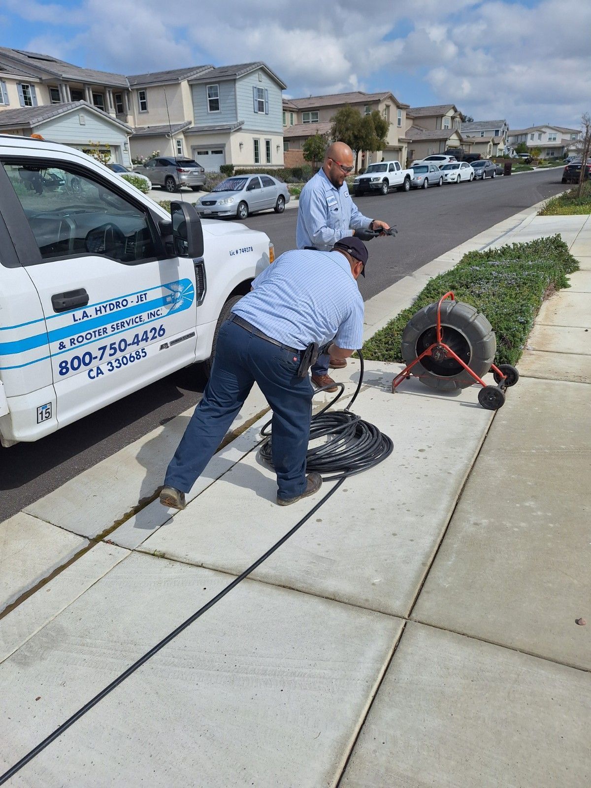 Two men inspecting a sewer line near a truck on a residential street. A camera reel is visible.