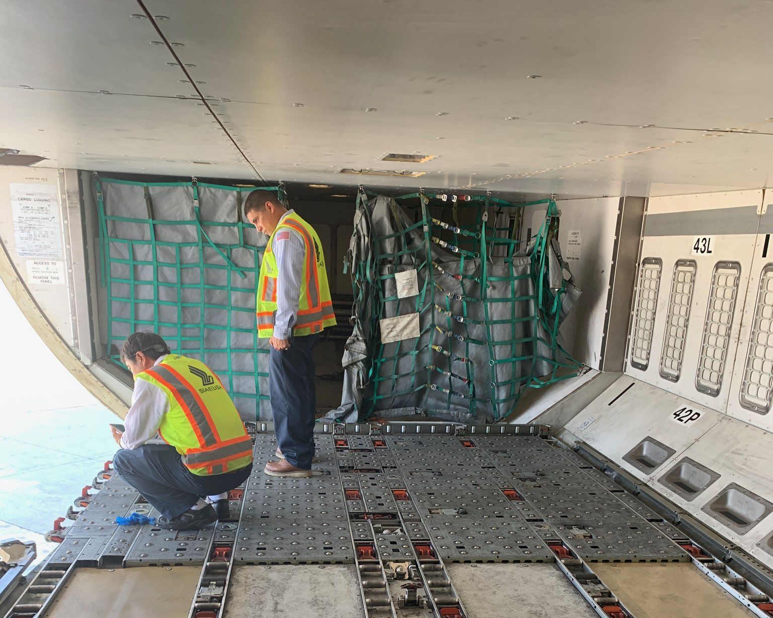 Two people in vests inspect cargo inside an airplane's hold.