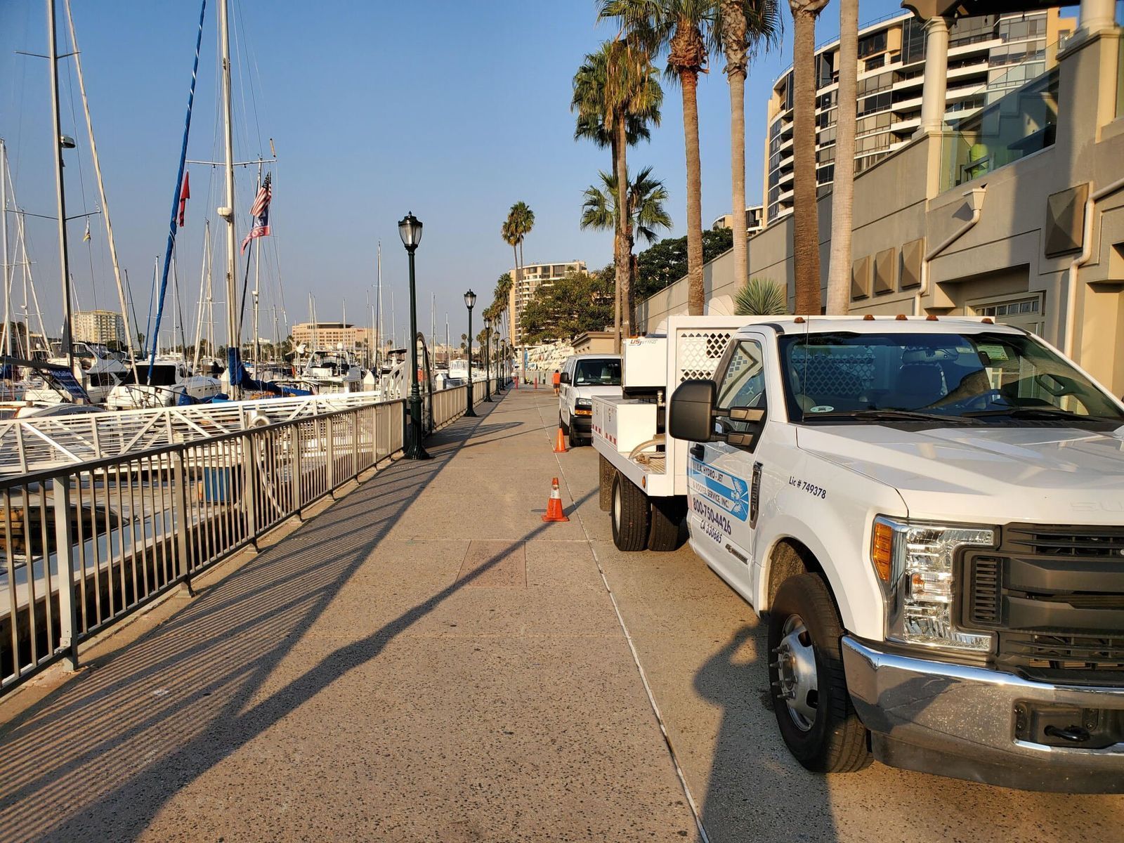 White work truck parked on a waterfront path with boats docked on one side and buildings on the other.