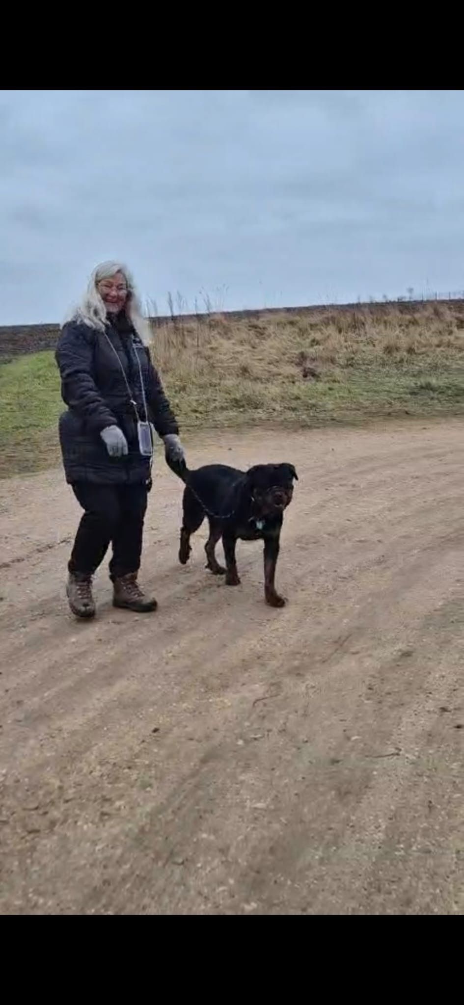 Woman walking a black dog on a dirt path under a cloudy sky.