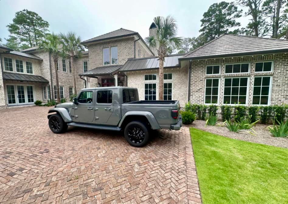 Gray Jeep truck parked in front of a large brick house on a brick driveway. Green grass on the side.