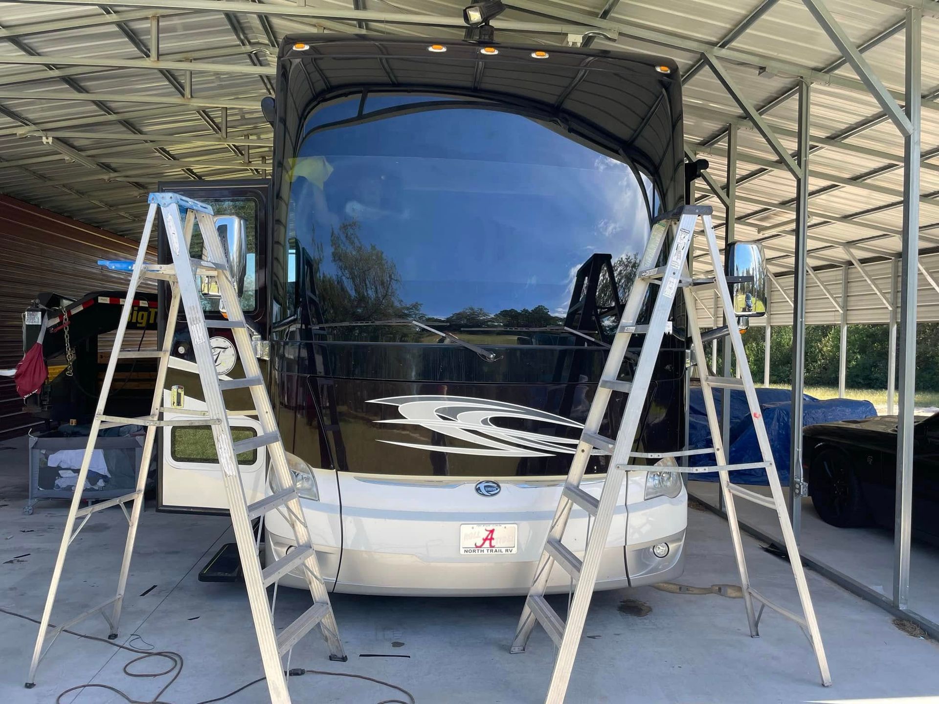 RV parked under a shelter with two ladders set up in front of it.