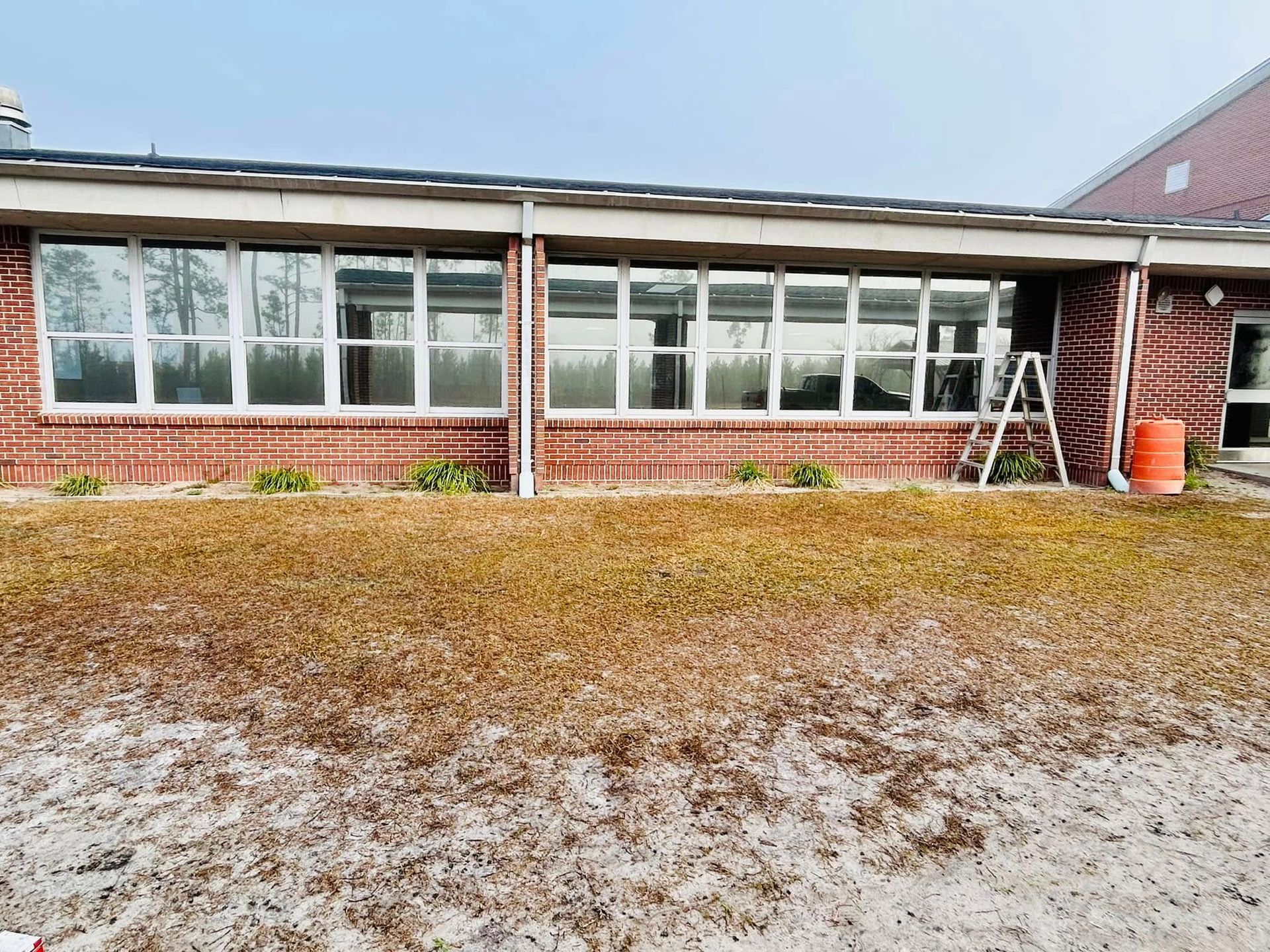 Brick building with large windows, ladder, and brown, patchy grass.
