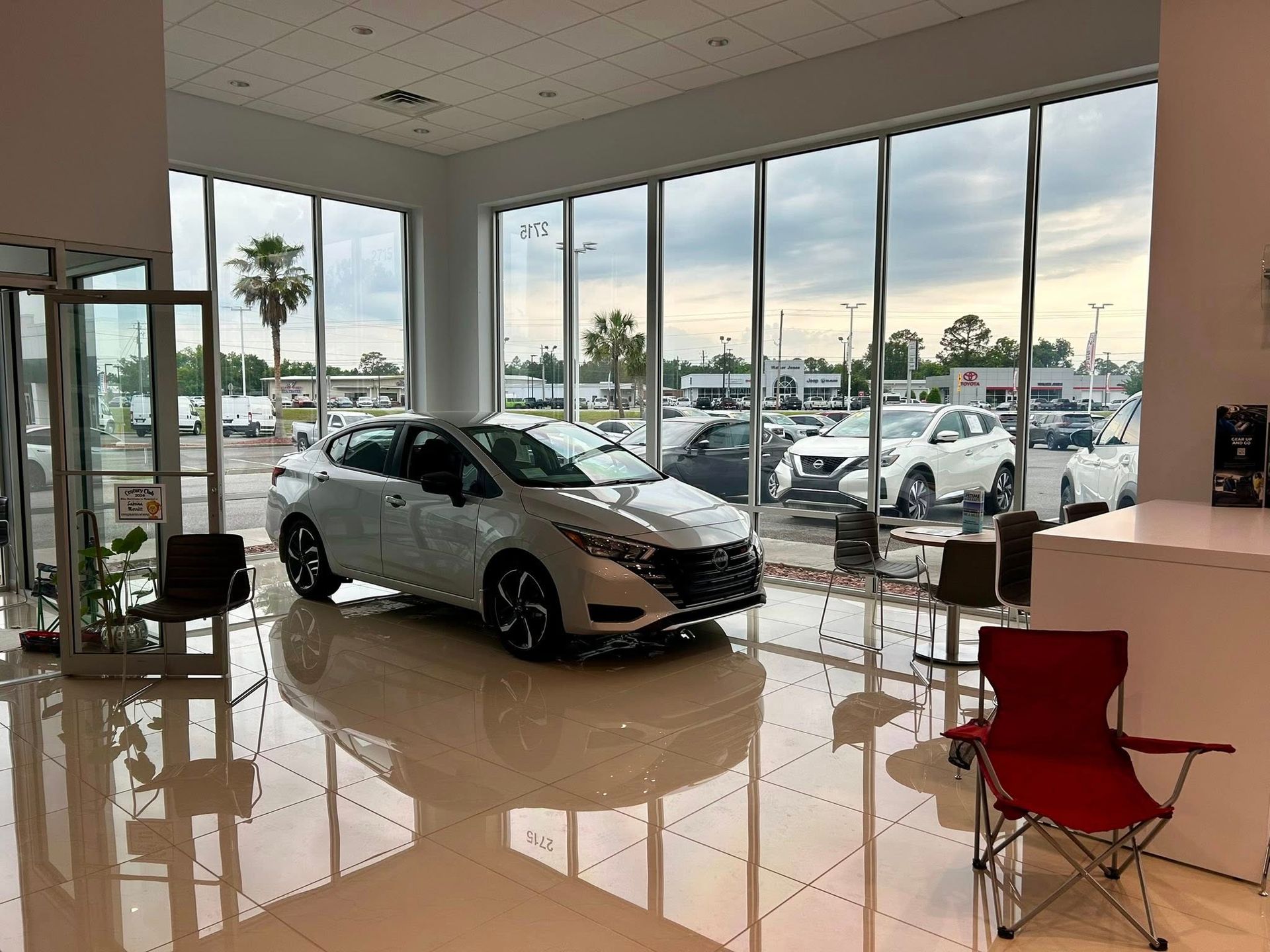 White car displayed in a car dealership showroom with large windows overlooking outdoor parking lot with other cars.
