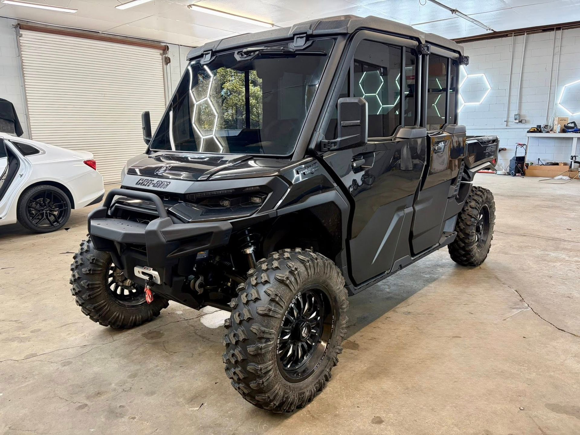 Black off-road vehicle with large tires, enclosed cabin, parked inside a garage, next to a white car.