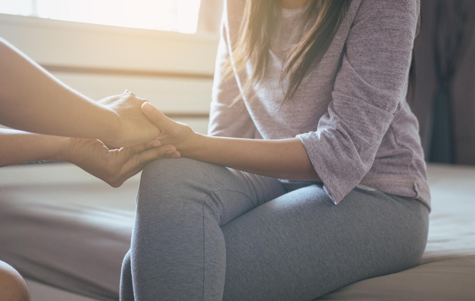 Person comforting another, holding hands, seated on a bed, sunlight streaming in.