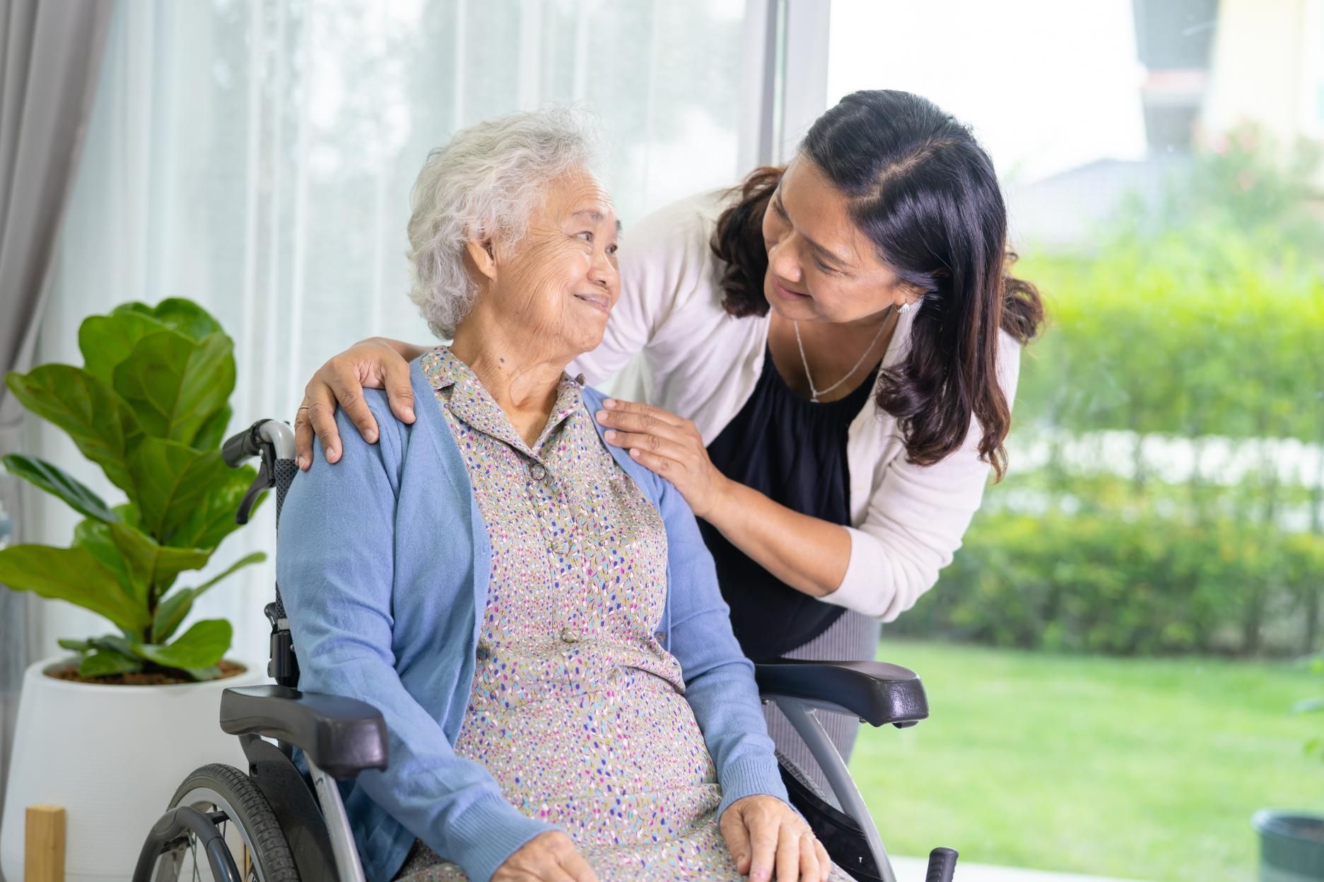 Woman in wheelchair with another woman's arm around her, smiling in front of a window with greenery.