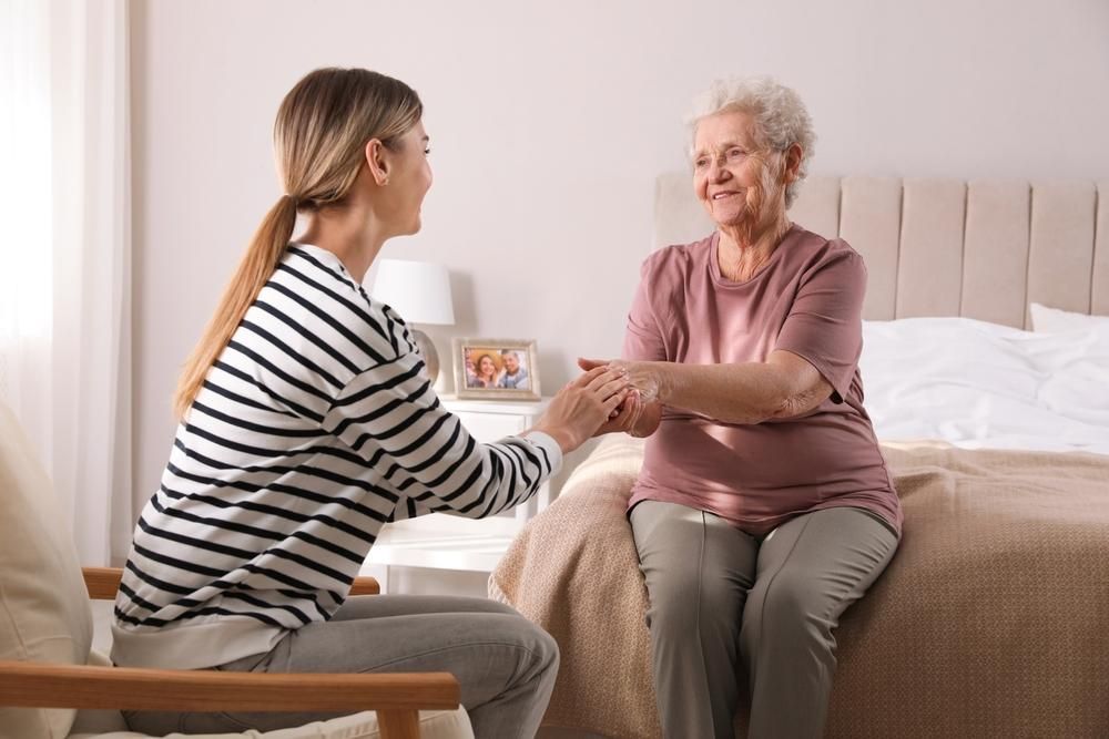 Woman helping elderly person sit up in a bedroom, smiling and holding hands.