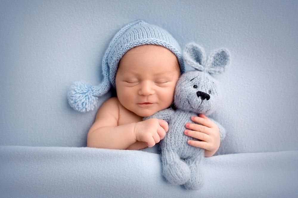 Baby wearing a blue knit hat, sleeping, holding a blue stuffed bunny, lying on a blue blanket.