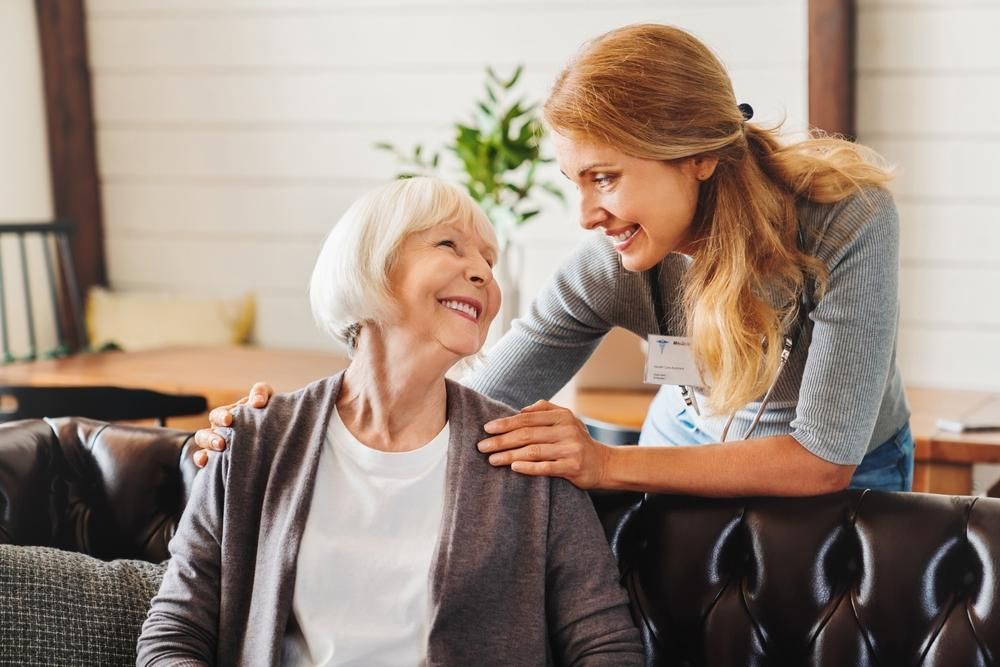 Woman smiles at older woman, arm on shoulder; indoor setting.