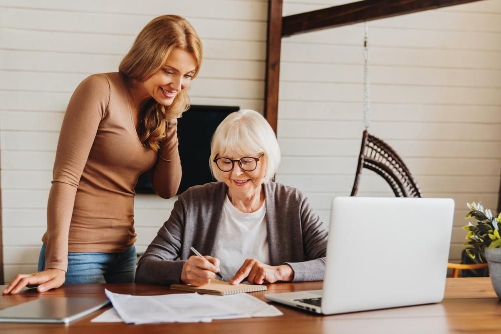 Woman helping senior woman with paperwork at a desk with a laptop; both smiling.