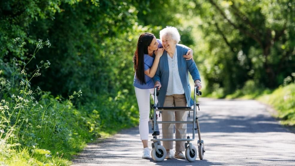 Woman assisting elderly person with a walker on a paved path outdoors.
