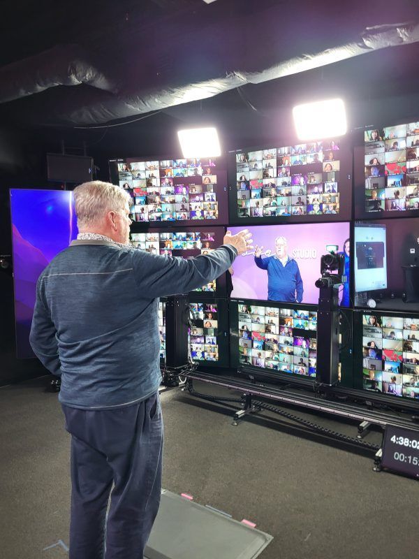 Man points at a wall of screens displaying a video conference in a studio. Lighting fixtures are above.