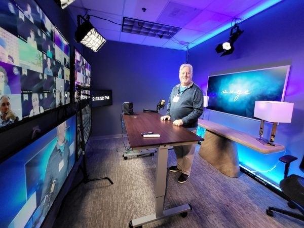 Man standing in a video studio with a large screen displaying an abstract image and multiple monitors. Blue lighting.