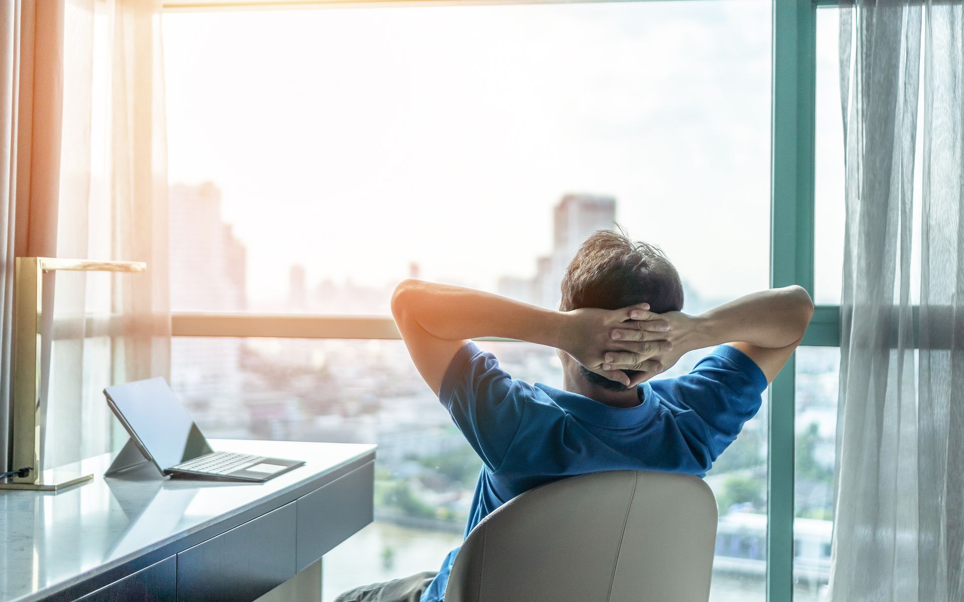 Person relaxing at desk, looking out large window with cityscape view. Hands behind head. Laptop visible.