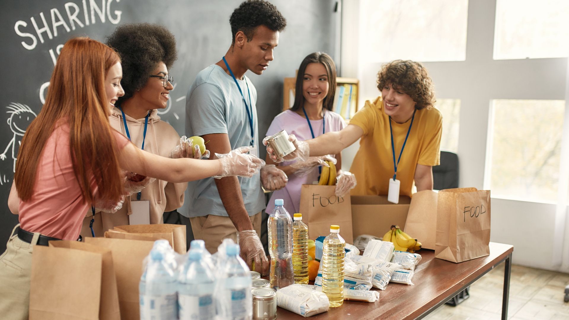 Volunteers packing food into bags at a table, including canned goods, bananas, and bottled water.