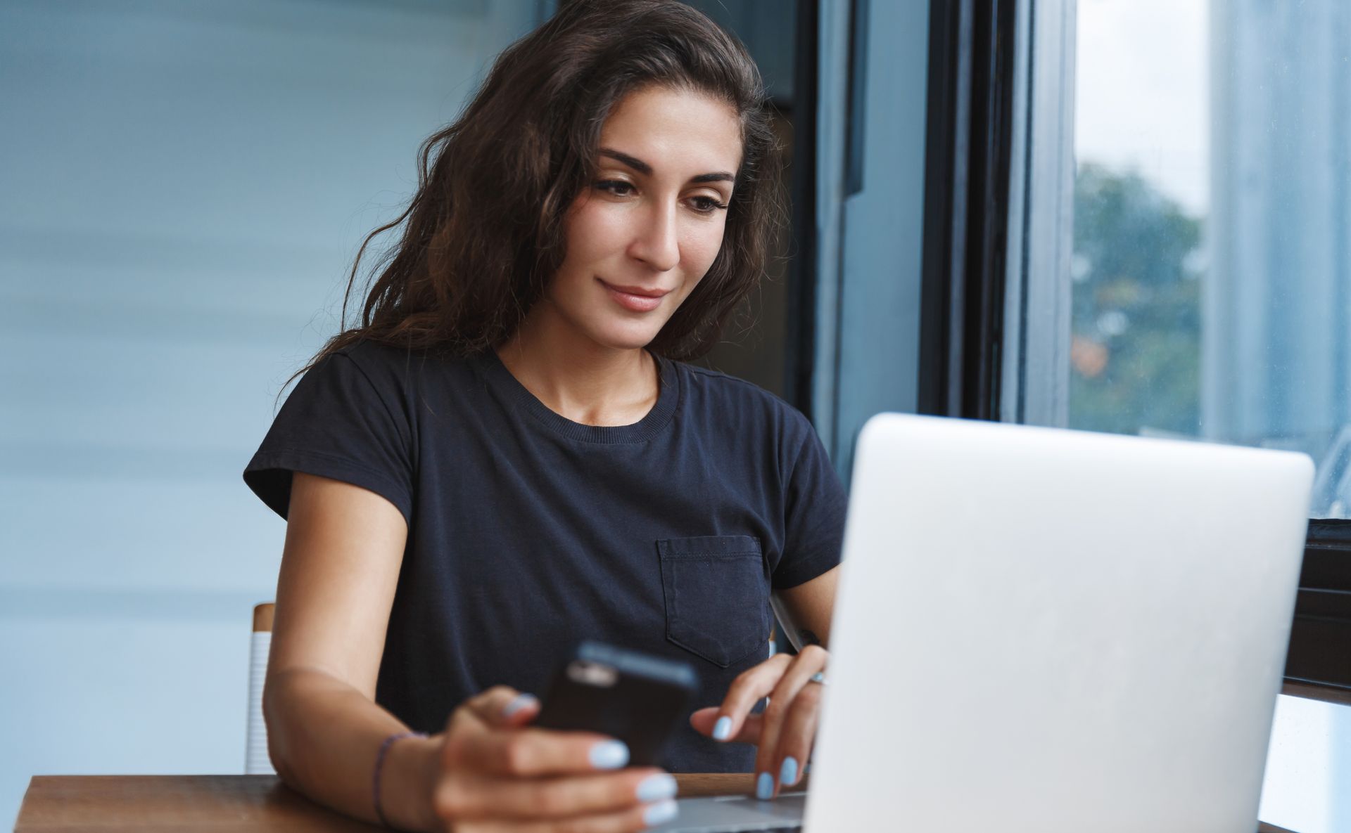 Woman using a laptop and a smartphone near a window; smiling, relaxed.